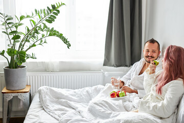Obraz na płótnie Canvas side view on cheerful couple have breakfast on bed at weekend, after waking up. they lie together on bed in bathrobes, eat healthy food