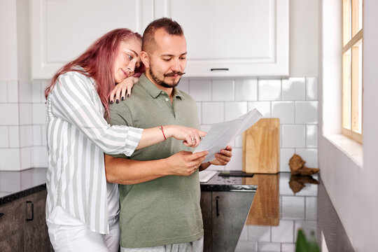man and woman with papers in the kitchen, young caucasian married couple checking bills documents in the kitchen, discuss