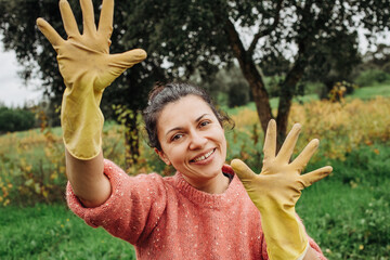 Woman in yellow rubber gloves dirty from work on the field showing open hands while smiling and standing at the field with grass and tree on background during autumn day