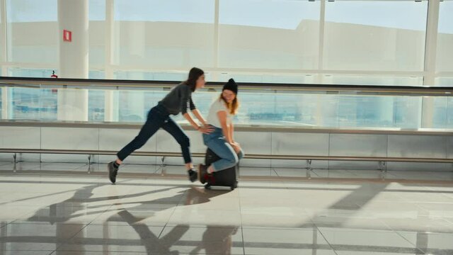 Two friends fool around, run and make jokes in airport. Friend push woman on top of suitcase, joking around in empty airport terminal, wait for delayed or cancelled flight. Teenagers travelling