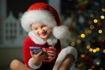 child a girl in a bright red suit and Santa hat laughing loudly tells friends on the phone about a gift for Christmas.