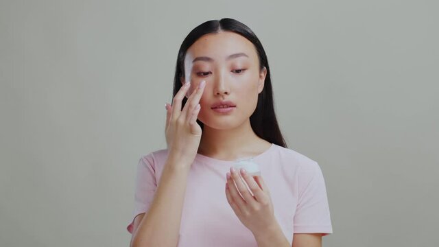 Young asian woman doing morning skincare routine applying face cream in front of mirror. Grey background. Beauty procedures. Selfcare.