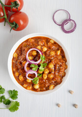 Top view of chole masala or chana local indian vegan food made of cooked chickpeas, tomatoes and cumin decorated with onion rings and parsley served in bowl on white wooden background. Vertical