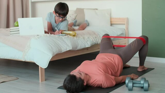 Home Isolation, Distance Learning, Remote Work, Fitness at Home, Online Training, Family Quarantine. Mom does physical exercises at home on the floor next to her son who does homework on the bed