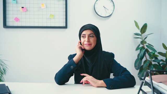 Smiling muslim businesswoman talking on smartphone near papers in office