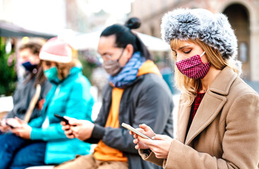 Blond girl using smart phone covered by mask on Covid second wave - New normal lifestyle concept with milenial people watching video on mobile smartphone - Vivid filter with focus on woman face