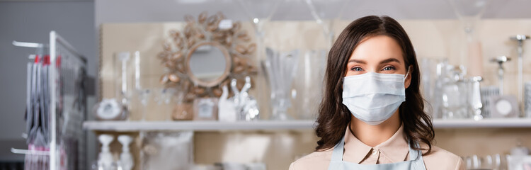  female florist in medical mask looking at camera with blurred rack of vases on background, banner