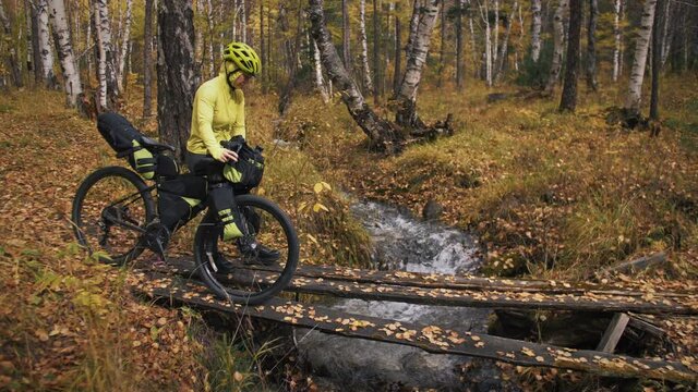 The woman travel on mixed terrain cycle touring with bikepacking. The traveler journey with bicycle bags. Sportswear in green black colors. The trip in magical autumn forest. Wooden bridge, river.