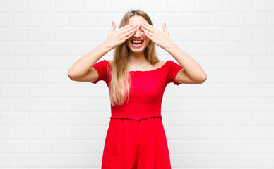 blonde woman smiling and feeling happy, covering eyes with both hands and waiting for unbelievable...
