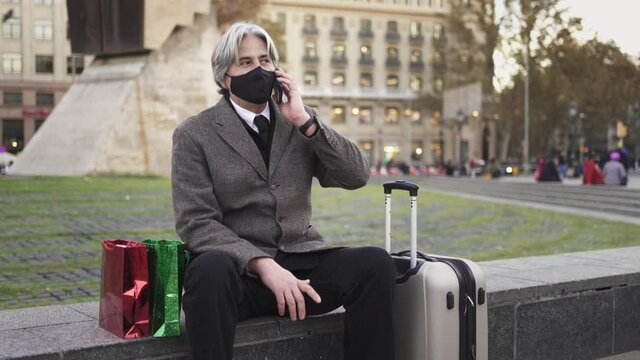 Man with face mask, presents and suitcase is going back home in Coronavirus Christmas season. Senior traveler talking on phone