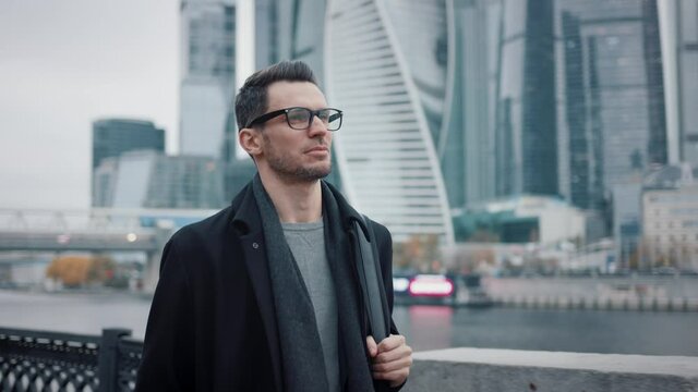 Man in black coat and eyeglasses walking on background of business skyscrapers in Moscow, slow motion. Middle shot of young man in the city business area
