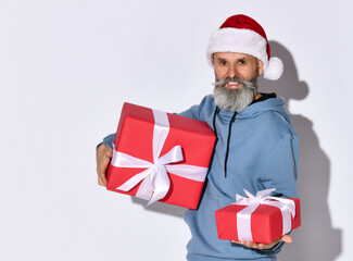 Stylish middle-aged man in Santa Claus hat holds out a gift to the camera, standing on a white background. Fashionable Santa wishes you a Merry Christmas and Happy New Year. Place for text.