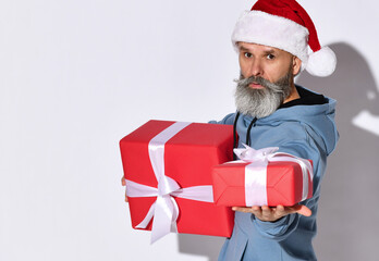 Stylish middle aged man in a Santa Claus hat holds out a gift to the camera while standing on a white background. Fashionable Santa wishes you a Merry Christmas and a Happy New Year. Place for text.
