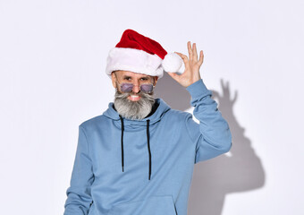 Fototapeta na wymiar Stylish senior man in a Christmas hat, glasses and a blue hoodie stands against a white background. Christmas sales and shopping concept. Space for text or promotional items.