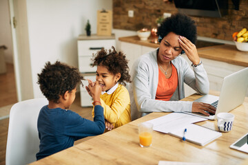 Black working mother using laptop and feeling stressed out while kids are playing beside her.