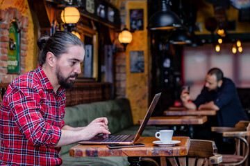 Portrait of a young bearded freelancer or blogger, sitting in a pub, working on a laptop computer. Other customer having a drink at an appropriate distance. Practicing social distancing. Remote work.