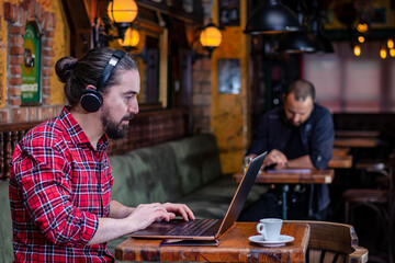Portrait of modern young bearded freelancer or blogger, sitting in a beer pub with earphones on his head, working on a laptop. Other customer working on a tablet computer at an appropriate distance. 