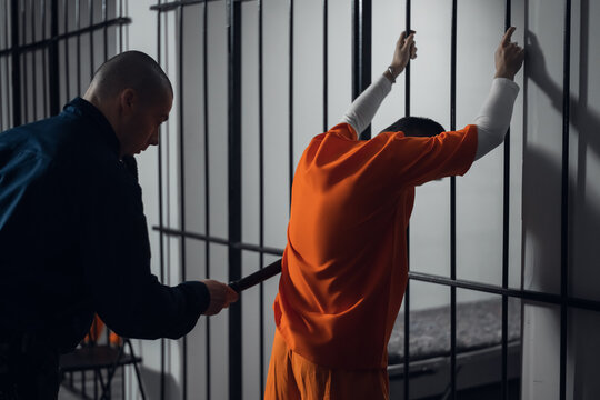 An armed guard searches a newly arrived criminal in a prison corridor against a backdrop of bars