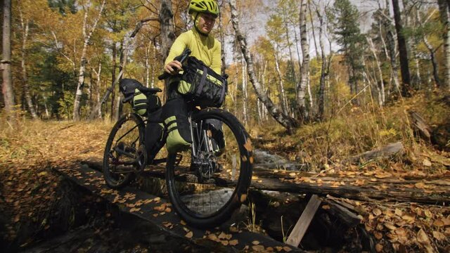 The woman travel on mixed terrain cycle touring with bikepacking. The traveler journey with bicycle bags. Sportswear in green black colors. The trip in magical autumn forest. Wooden bridge, river.