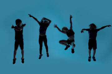 Silhouetted full length shot of four little sportive kids looking joyful while posing, jumping isolated over blue background