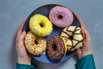 Two hands holding plate of colourful sugary round glazed donuts with colored sprinkles on gray background top view