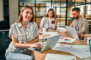 Glad to be on the team. Beautiful cheerful woman with a laptop with a smile looks into the camera, sitting at an office table with her colleagues.