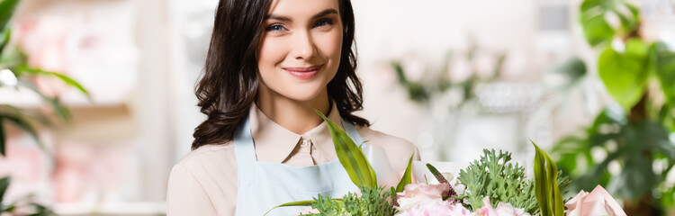 young florist smiling at camera while holding bouquet in flower shop on blurred background, banner
