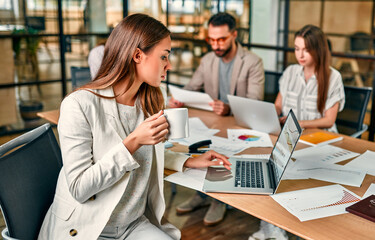 Beautiful caucasian business woman with a cup of coffee or tea working at a laptop sitting at an office table with her colleagues.