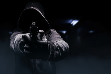 Photo of a creepy horror criminal in black hoodie aiming and attacking revolver gun.