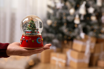 Man holding snow globe with Christmas tree at home, closeup. Space for text