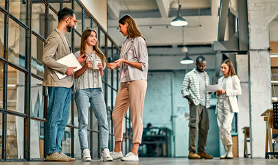 Obraz premium A group of young colleagues are discussing work while standing in the office hallway. A successful hipster team in a coworking space. Freelancers.