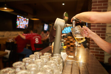 The bartender pours a beer in a beer cups with football fans in the background. Draught beer.