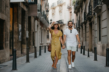 A girl in a hat and a yellow dress with a plunging neckline and her boyfriend with a beard are walking holding each other's hand in old Spain town. A couple of tourists on a date in Valencia.