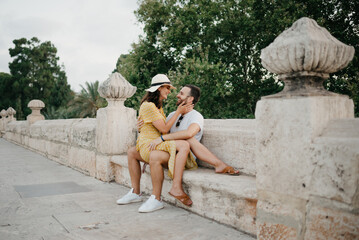 A girl in a hat and a yellow dress with a plunging neckline is sitting on the legs of her boyfriend with a beard on the ancient bridge in old Spain town. A couple of tourists on a date in Valencia.