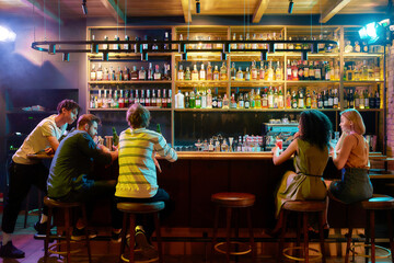 Rear view of three guys drinking beer, looking at women, two girlfriends sitting at the bar counter. Friends spending time at night club, restaurant - Powered by Adobe
