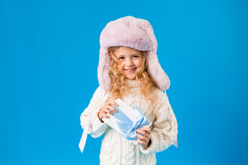 Cute little blonde girl in a pink hat and white knitted sweater smiles and holds a gift box on a blue background. Space for text