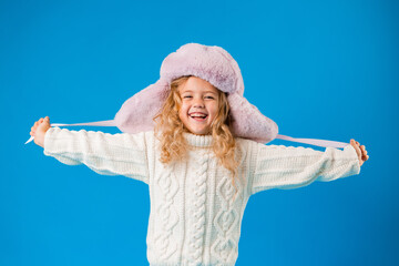 portrait of a little blonde girl in a winter hat and knitted sweater on a blue background. baby girl in winter clothes smiling on blue background in Studio, space for text
