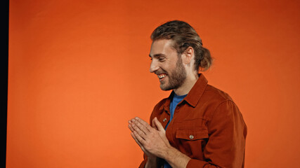 positive young and bearded man smiling and standing with praying hands on orange
