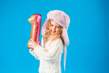 little girl in winter clothes smiles and holds a balloon in the shape of a number one on a blue background