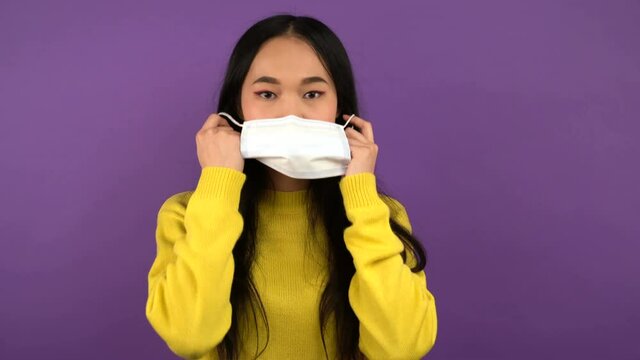 young Asian woman takes off her protective medical mask. lady in yellow sweater on purple isolated background. 4K
