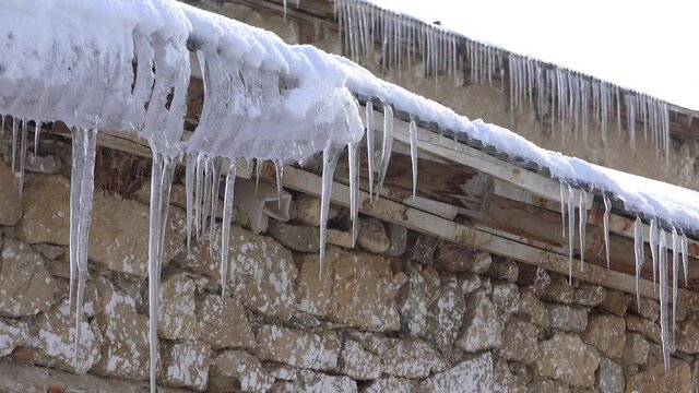 Ice eaves hanging from the corrugated aluminum sheet roof of the village house in winter. Snowy stone wall villages houses in the heavy continental climate.Icicles can form wherever water seeps out 4K