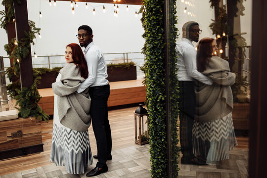 Beautiful couple stand at the balcony, handsome African American man hug gorgeous white woman wrapped in a blanket, enjoy tender moments, winter holidays, Christmas morning concept