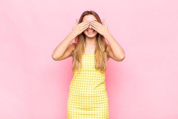 blonde woman smiling and feeling happy, covering eyes with both hands and waiting for unbelievable...