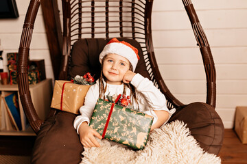 Happy cute child sitting on chair with present. Studio shot of little birthday girl wears cute dress and santa hat waiting for Christmas and New Year