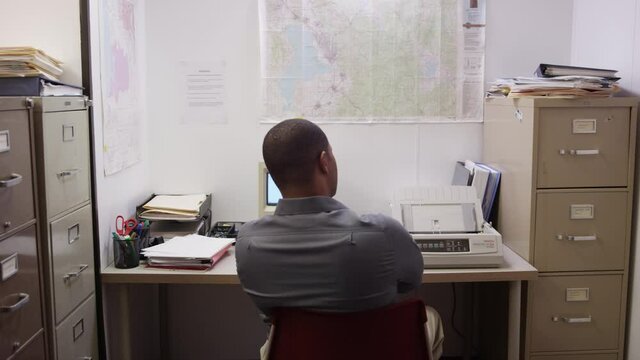 Black man waits for printer to start printing in small cluttered office