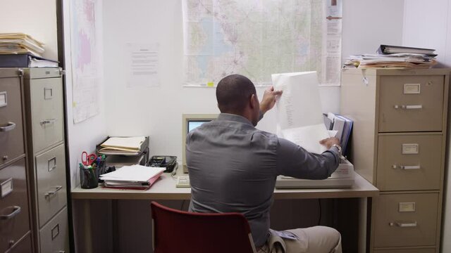 Black man waits for document to finish printing in small cluttered office