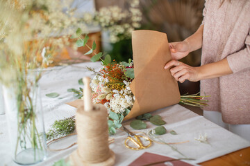 hand of florist wrapping a bouquet of dry flowers, eucalyptus in craft paper in flower shop, green,...