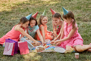 Fototapeta premium Little girls celebrate their birthday in the summer on a picnic. They eat sweets. A lot of gifts in festive bright packages.