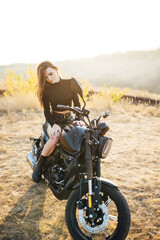 Beautiful girl in black clothes sits on motorcycle and poses for camera. Evening sunset