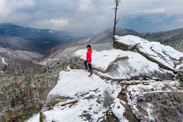 Aerial view on a runner girl in a red sweatshirt stands on a snowy rock admiring the beauty of the mountains on a cloudy day.